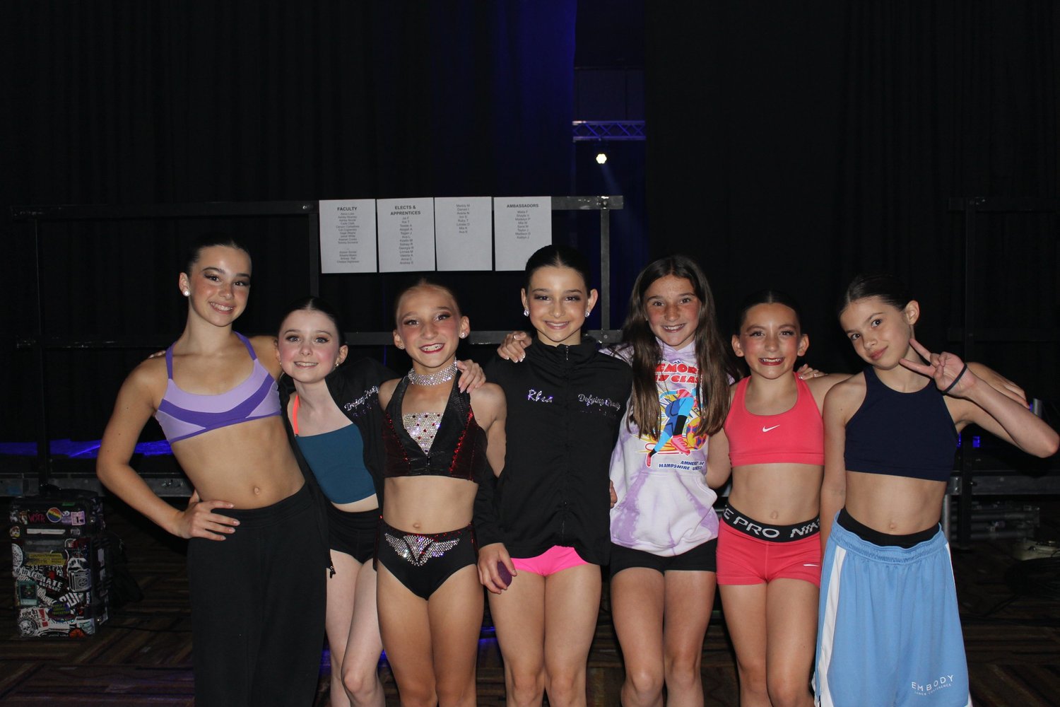 Seven young dancers smiling together backstage at the competition.